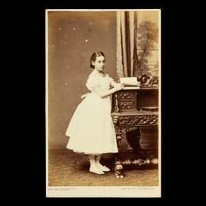 Studio portrait of a girl in white, turning the pages of a photograph album. Melbourne, circa 1870.