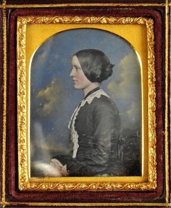 Tinted daguerreotype portrait of a seated woman in profile with painted cloud backdrop, dated February 1854.