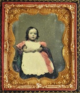 Ambrotype of a young girl seated in a chair, her hands clasped. Early 1860s.
