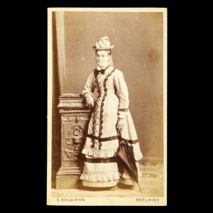 Studio portrait of a woman in summer dress, wearing a fancy hat decorated with flowers and holding a parasol. Adelaide, late 1870s.