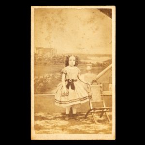 Studio portrait of a young girl posed before a painted backdrop with a view of St. Mary's Cathedral and Sydney Harbour. Sydney, early 1868.