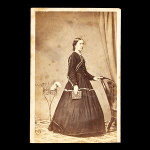 Studio portrait of a young woman holding a carte de visite album, her white gloves placed in a wire "glove basket". Ballarat, Victorian goldfields, c.1865.