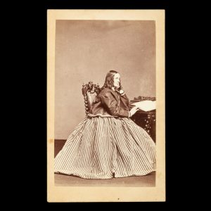 Profile portrait of a woman with bottle curls and wearing a striped dress, seated at a writing desk with her hand on an open book. Adelaide, mid 1860s.