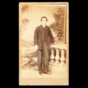 Studio portrait of a young man posed in front of a painted backdrop with a subtropical landscape. Sydney, c.1865.